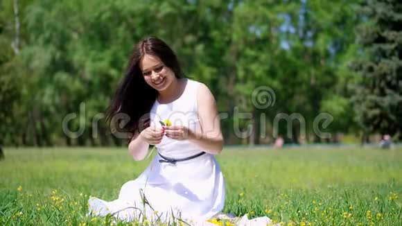 夏天美丽的黑发女人穿着白色连衣裙坐在绿色的草坪上草地上编织着黄色蒲公英的花环微笑着视频的预览图