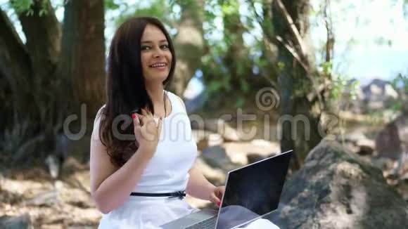 美丽的黑发浪漫女人穿着白色连衣裙坐在石头上在户外的笔记本电脑上工作她在博客上做文章视频的预览图