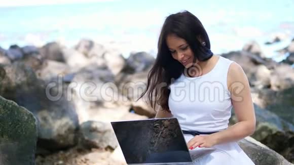 美丽的黑发女商人穿着白色连衣裙坐在石头上在户外工作在笔记本电脑上躺着视频的预览图