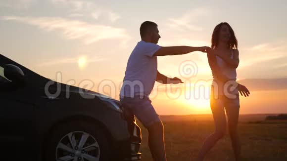 幸福夫妇的剪影停留在新车的天空下日落时分美丽的年轻夫妇站在汽车附近视频的预览图