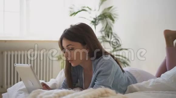 年轻女子在卧室里躺在床上玩平板电脑视频的预览图