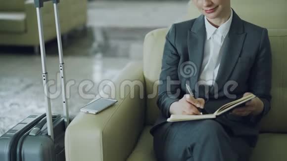 漂亮的女商人坐在酒店大堂的扶手椅上微笑着写着记事本望着一边视频的预览图
