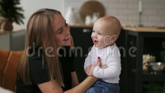 妈妈抱着宝宝孩子微笑着看着心爱的妈妈圣诞节时一起站在白色的厨房里视频的预览图
