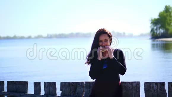 夏天靠近河滩美丽的黑发女人的肖像留着长长的黑发她正在吃快餐汉堡包视频的预览图
