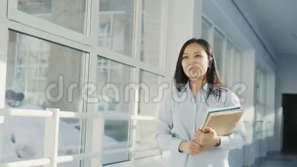 年轻美丽的亚洲女孩走在大学走廊上手里拿着课本和报纸注意到朋友挥手示意他们视频的预览图