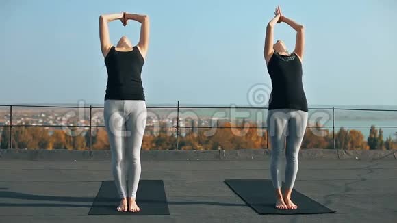 两个正在练习瑜伽体式的年轻女性举起手臂在屋顶上摆姿势户外视频的预览图