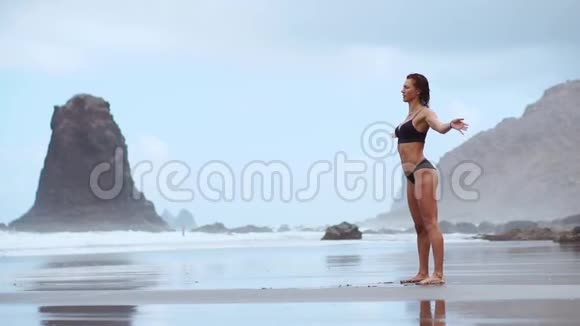 一位美丽的女子在海滩上用黑色的火山沙做体操练习健康的身体视频的预览图