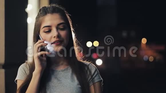 年轻漂亮的女人在她的智能手机上说话晚上照亮了街道智能手机科技概念视频的预览图