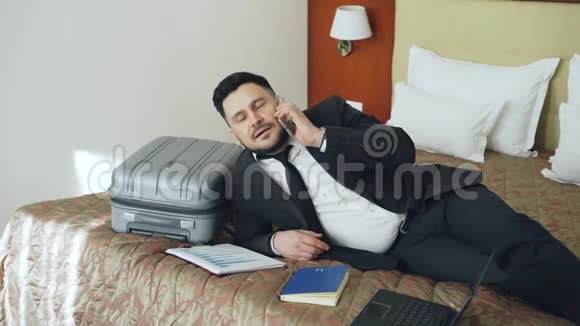 开朗的商人在酒店房间里躺在床上谈论手机旅游商业和人的概念视频的预览图