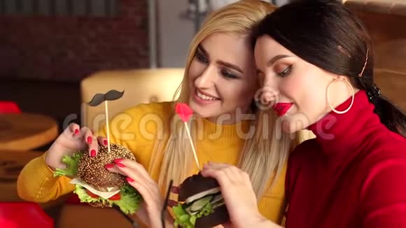 两个美丽的年轻女孩化妆鲜艳在咖啡馆里吃汉堡慢动作视频的预览图