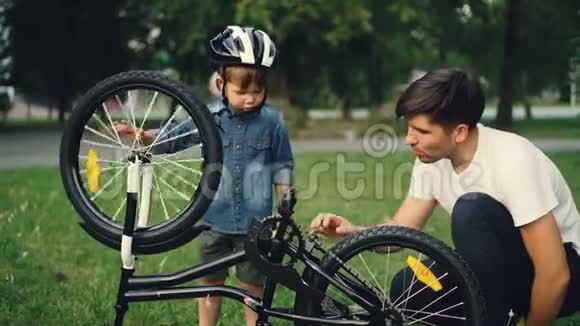 小男孩正在旋转自行车和踏板而他的父亲正在公园的草坪上和他说话家庭视频的预览图