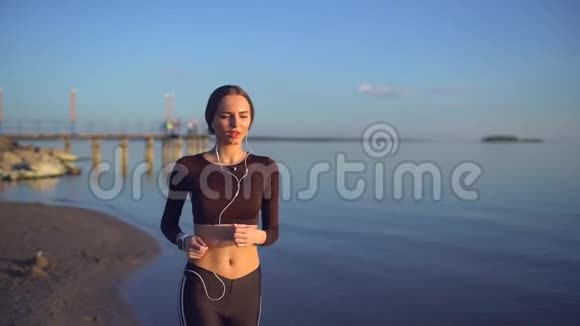 带着智能手表在海上慢跑的年轻女人年轻的运动女孩在日落时在海滩上使用智能手表太阳透镜耀斑视频的预览图