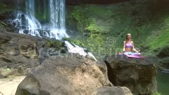 在公园瀑布旁金发姑娘坐在岩石上的莲花姿态视频的预览图