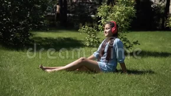 年轻女孩在阳光明媚的天气里坐在公园的草地上用智能手机听耳机里的音乐4K视频的预览图