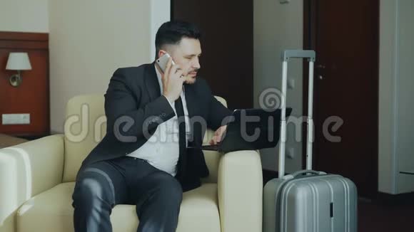 自信的年轻商人穿着西装坐在扶手椅上看着笔记本电脑一边用手机说话视频的预览图