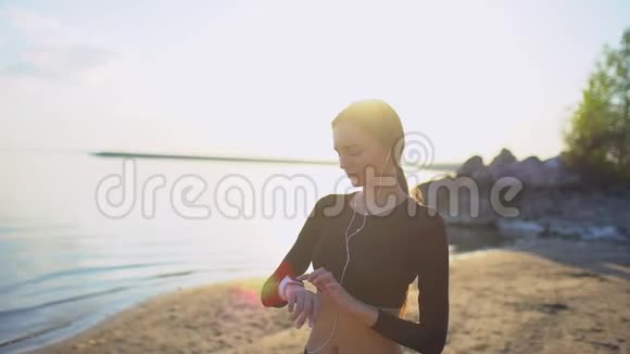 带着智能手表在海上慢跑的年轻女人年轻的运动女孩在日落时在海滩上使用智能手表太阳透镜耀斑视频的预览图