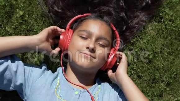 漂亮的黑发女孩躺在公园的草地上戴着红色耳机听音乐拥有一个好的视频的预览图