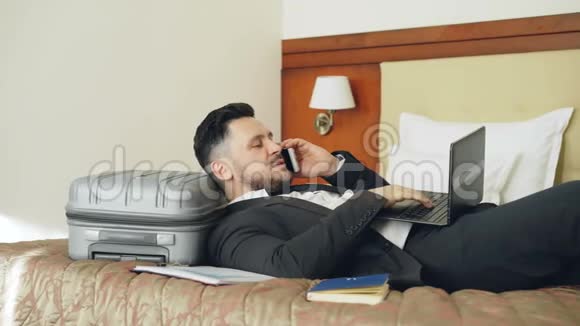 年轻的商人躺在床上和手提箱里讲电话用他酒店房间里的笔记本电脑工作旅行商务视频的预览图