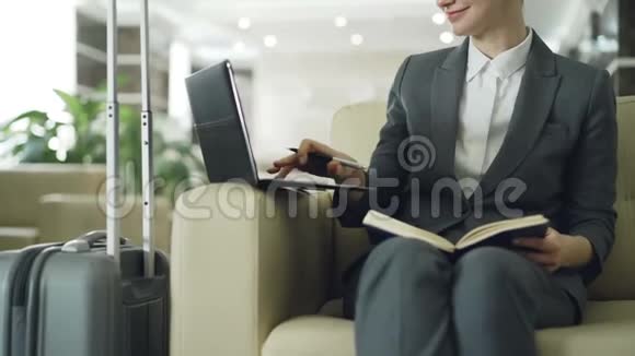 金发女商人坐在酒店大堂的扶手椅上用记事本和笔记本电脑写字视频的预览图