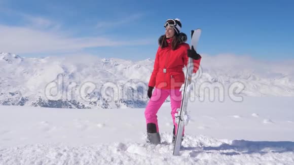 妇女手持滑雪板戴着护目镜站在白雪皑皑的山脉背景下视频的预览图