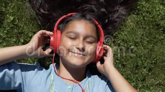美丽的女孩留着长长的黑发躺在公园的草地上听着红色耳机上的音乐视频的预览图