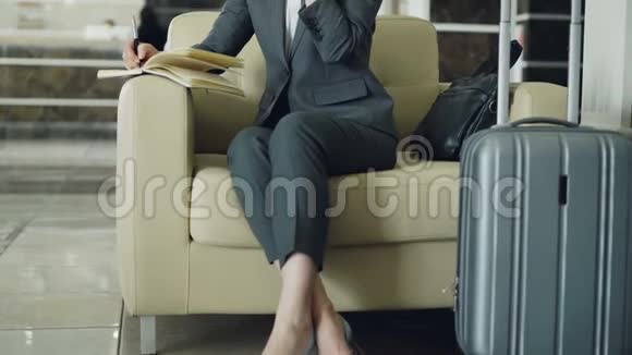 坐在酒店大堂扶手椅上的金发开朗的女商人倾斜着在大厅里讲手机和写笔记视频的预览图