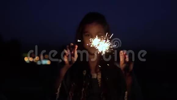 一个留着深色长发的年轻女孩在城市的背景下晚上拿着烟火高兴起来慢动作高清高清视频的预览图