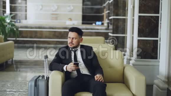 潘是一位体贴的商人他一边看一边用智能手机坐在豪华酒店的扶手椅上带着行李视频的预览图