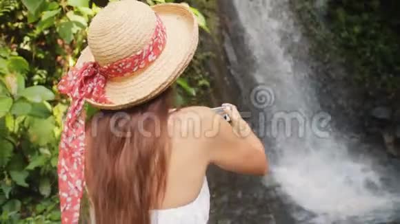 年轻的快乐旅游女孩穿着白色连衣裙和草帽制作照片惊人的丛林瀑布使用移动智能手机自然视频的预览图