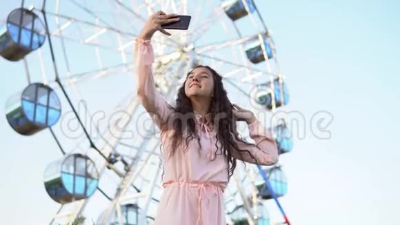 一个穿着连衣裙留着长发的女孩用智能手机在摩天轮附近自拍4K视频的预览图