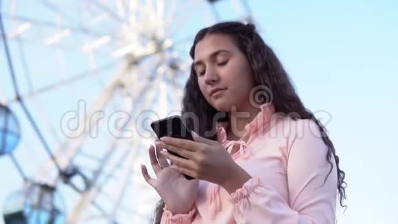 这个女孩正在使用一部智能手机站在大摩天轮附近慢动作视频的预览图