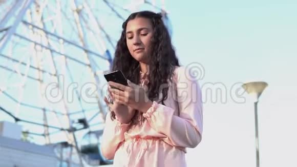 一个长发女孩使用智能手机站在大摩天轮和游乐园附近慢动作视频的预览图