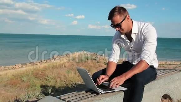 穿着白色衬衫和太阳镜的商人在海滨带着笔记本电脑背景是大海风疹鸡肉视频的预览图