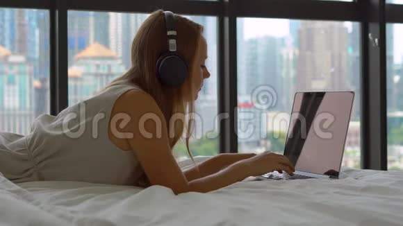 躺在床上的年轻女性自由职业者使用无线耳机听音乐并在笔记本电脑上工作视频的预览图