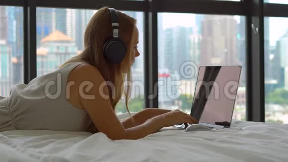 躺在床上的年轻女性自由职业者使用无线耳机听音乐并在笔记本电脑上工作视频的预览图