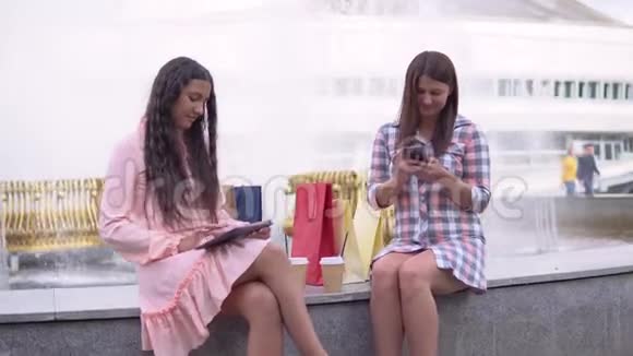 购物后的两个女孩正坐在喷泉附近的公园里用手机和平板电脑玩得很开心4K视频的预览图
