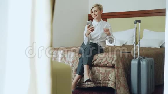 幸福的女商人通过在线视频聊天与家人交谈使用智能手机摄像头坐在酒店房间的床上视频的预览图