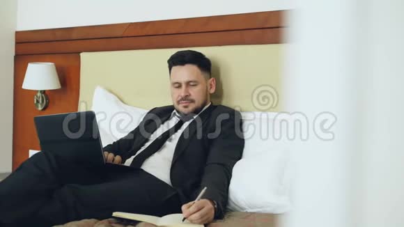 自信的商人躺在床上在酒店房间工作时用笔记本电脑和记事本写笔记视频的预览图