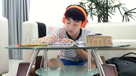 亚洲男孩戴着耳机在客厅的桌子上做作业视频的预览图