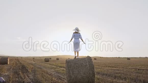 后景年轻女孩穿着连衣裙戴着大帽子站在干草堆上伸出双手在田野的夕阳下玩得很开心视频的预览图