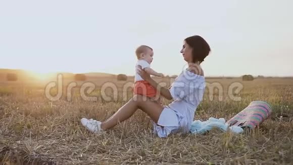 一个穿着浅色连衣裙的年轻女子正坐在田野的草地上和一个可爱的婴儿玩耍妈妈和儿子快乐视频的预览图