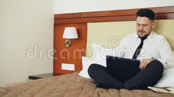 微笑的商人放下笔记本电脑在酒店房间里放松地坐在床上旅费视频的预览图