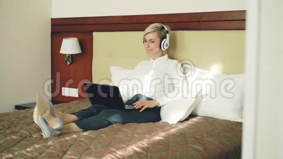 快乐的女商人戴着耳机听音乐微笑着跳舞唱歌同时使用笔记本电脑躺在床上视频的预览图