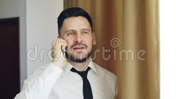穿着白色衬衫和领带站在酒店房间窗户附近的成人微笑商人用手机交谈视频的预览图