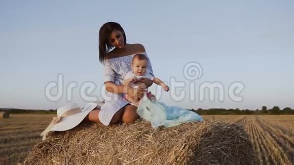 一个漂亮的女孩和小儿子坐在干草堆上的田野里和他玩穿蓝色衣服的漂亮年轻女子视频的预览图