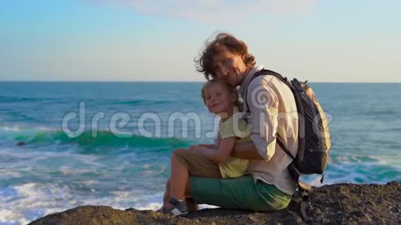 一对父子坐在岩石上看着塔纳罗得寺附近的海浪慢镜头视频的预览图