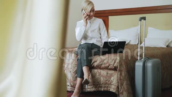 一位穿着白色衬衫的微笑女商人潘在酒店坐在床上用笔记本电脑和手机聊天视频的预览图
