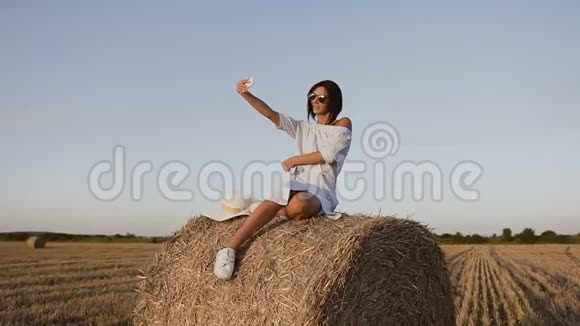漂亮的年轻女人穿着蓝色的衣服和太阳镜在干草堆上享受大自然在电话里拍了一张照片视频的预览图