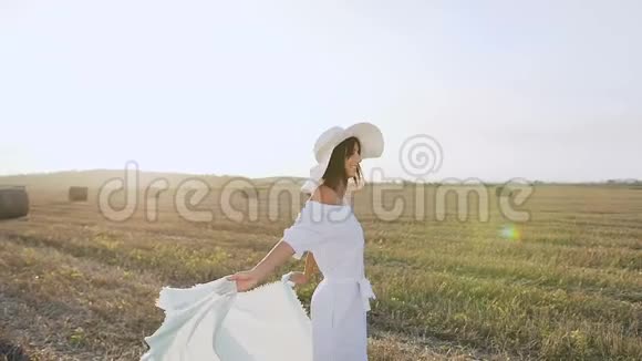 迷人的性感女孩穿着一件长裙子戴着围巾的大帽子在日落的夏天在田野里旋转美容视频的预览图