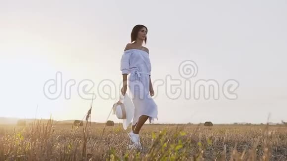 后景日落时分一个年轻的黑发女人在田野里散步穿着带帽子的蓝色长裙的漂亮女孩视频的预览图